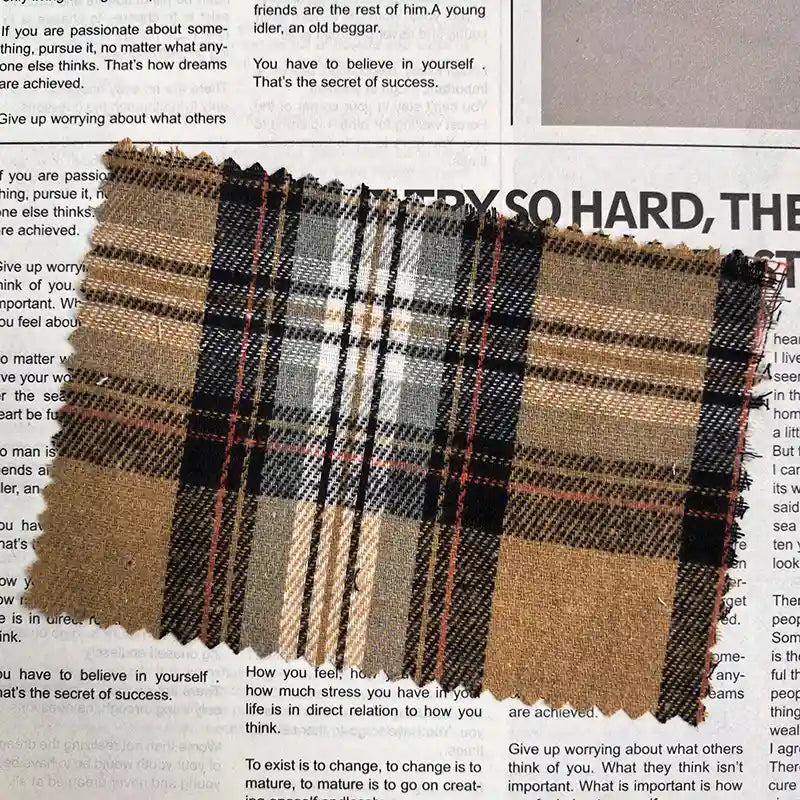 A piece of Longan Craft’s Brown Plaid Cotton Flannel Fabric By The Yard, featuring a retro pattern in brown, black, white, and red tones, rests atop an open magazine with visible text.