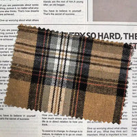 A piece of Longan Craft’s Brown Plaid Cotton Flannel Fabric By The Yard, featuring a retro pattern in brown, black, white, and red tones, rests atop an open magazine with visible text.