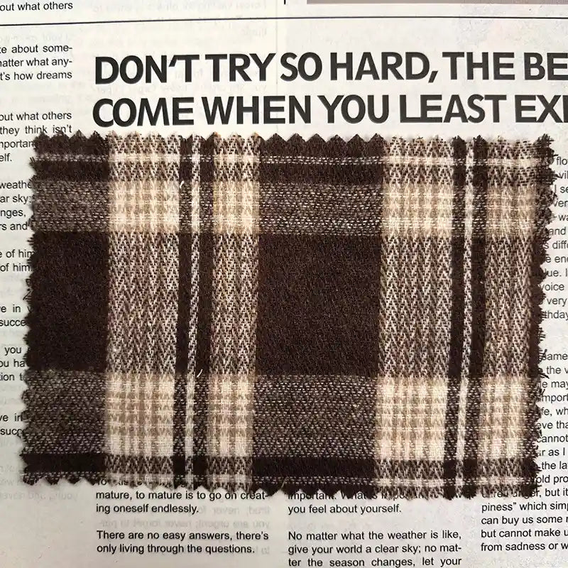 A swatch of Longan Craft’s Brown Plaid Cotton Flannel Fabric by the yard, a lightweight polyester brushed flannel material ideal for casual pants, rests atop a newspaper featuring bold black headlines and smaller print beneath.