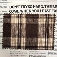 A swatch of Longan Craft’s Brown Plaid Cotton Flannel Fabric by the yard, a lightweight polyester brushed flannel material ideal for casual pants, rests atop a newspaper featuring bold black headlines and smaller print beneath.