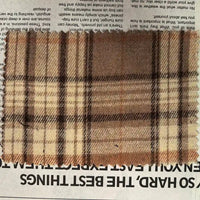 A rectangular piece of Longan Craft’s Brown Plaid Cotton Flannel Fabric By The Yard, a lightweight polyester brushed flannel material in beige and brown plaid, lies on top of a newspaper with its edges cut in a zigzag pattern.