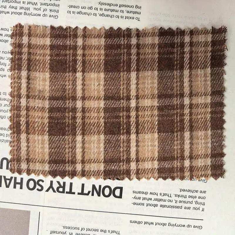 A swatch of Longan Craft’s Brown Plaid Cotton Flannel Fabric By The Yard—made from lightweight polyester brushed flannel—rests with its zigzag-cut edges atop a newspaper, allowing some print to show through beneath.