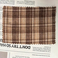 A swatch of Longan Craft’s Brown Plaid Cotton Flannel Fabric By The Yard—made from lightweight polyester brushed flannel—rests with its zigzag-cut edges atop a newspaper, allowing some print to show through beneath.
