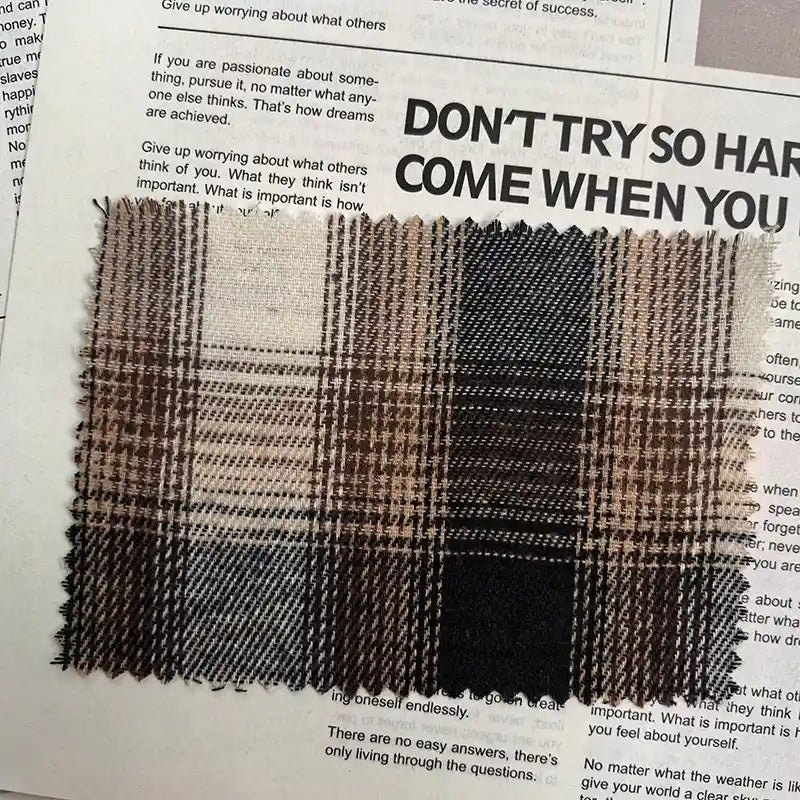 A swatch of Longan Craft’s Brown Plaid Cotton Flannel Fabric by the yard, featuring a retro brown, beige, and black plaid pattern with zigzag-cut edges, rests atop a newspaper page with bold and fine printed text in the background.