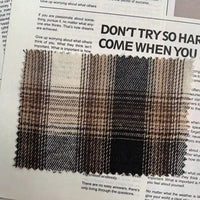 A swatch of Longan Craft’s Brown Plaid Cotton Flannel Fabric by the yard, featuring a retro brown, beige, and black plaid pattern with zigzag-cut edges, rests atop a newspaper page with bold and fine printed text in the background.