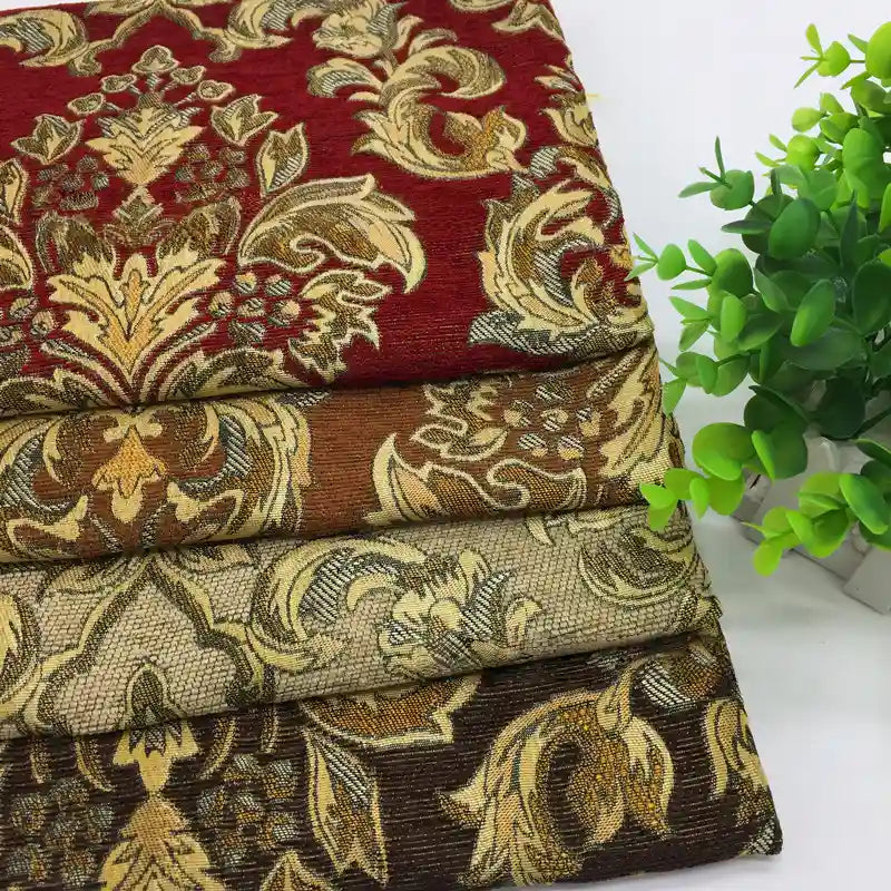 Four folded Chenille Damask Fabrics by Longan Craft in red, brown, beige, and dark brown with ornate floral jacquard patterns are stacked beside a green potted plant—ideal upholstery or décor fabric by the yard.