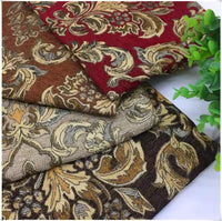 Three folded pieces of Longan Craft's Chenille Damask Fabric By The Yard in red, brown, and beige are stacked together with a leafy plant nearby—an ideal floral jacquard damask fabric for elegant upholstery or home décor projects.