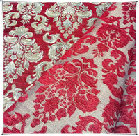 A close-up of Longan Craft’s Chenille Damask Fabric by the Yard shows its ornate red and gold floral jacquard pattern. A section is folded, revealing the reverse side with a muted red and beige design—perfect for upholstery projects.