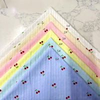 Six cotton dobby fabric swatches from Longan Craft’s Cherry Printed Textured Dobby collection (white, cream, pink, yellow, light blue, blue) are fanned on marble. Each features cute cherries and is labeled 1#–6#.