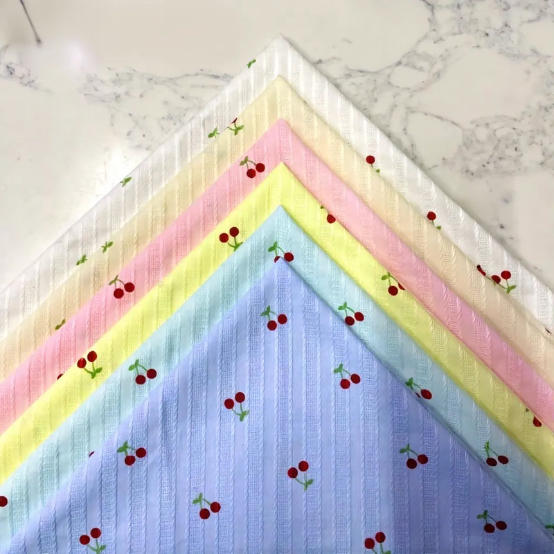 Five pastel cotton squares—white, yellow, pink, green, and blue—from Longan Craft's Cherry Printed Textured Dobby Fabric are stacked in a fan shape. Each features ribbed stripes and a small repeating cherry pattern.