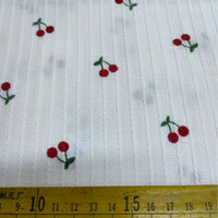 Cherry Printed Textured Dobby Fabric by Longan Craft features small red cherries and green stems on a striped cotton dobby weave, perfect for apparel. Shown next to a yellow centimeter-marked tape for scale.