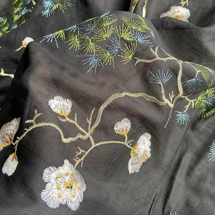 A close-up of Longan Craft’s Chinoiserie Embroidered Silk Fabric By The Yard, showcasing intricate white flowers with yellow centers and green pine needles on sheer black premium mulberry silk-cotton blend.