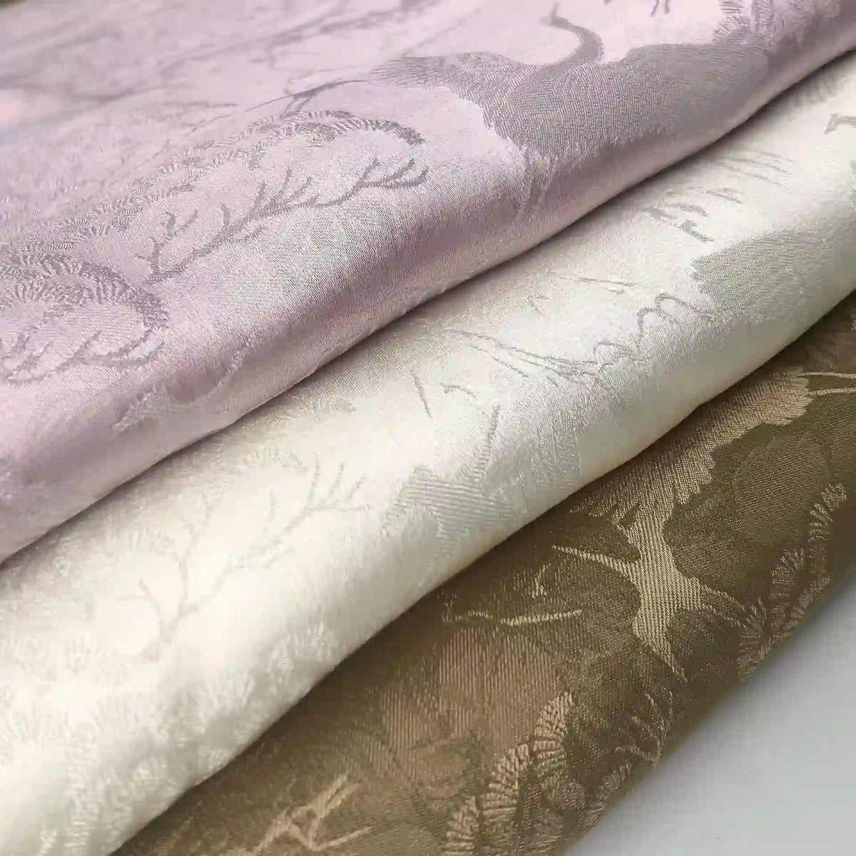 Three folded pieces of Longan Craft Chinoiserie Polyester Viscose Fabric in pastel pink, off-white, and taupe, each with a subtle shiny botanical jacquard pattern. Ideal for curtains and inspired by Chinoiserie designs.