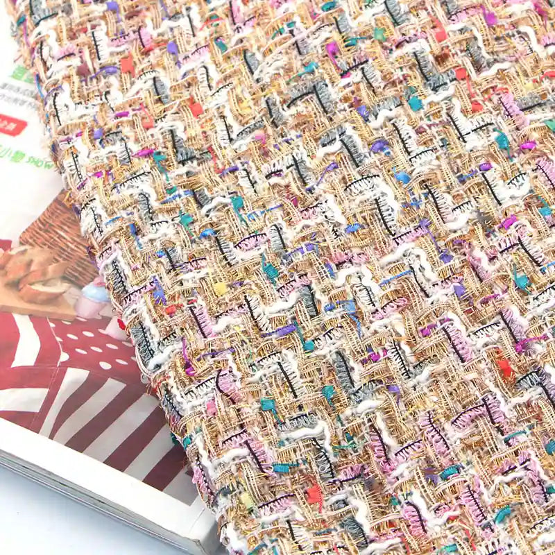 Close-up of the Longan Craft Colorful Boucle Tweed Fabric By The Yard, featuring thick, textured weaving with vibrant gold, pink, blue, and white threads. This luxury tweed material for blazers is displayed atop magazines—one showing a red and white striped cover with a wicker basket image.