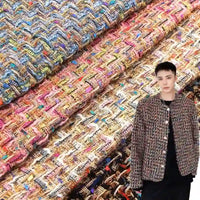 A person wearing a jacket crafted from Longan Craft’s Colorful Boucle Tweed Fabric By The Yard—a thick, textured woven tweed material—stands in front of stacked rolls of the luxury tweed fabric for blazers in various bright hues.