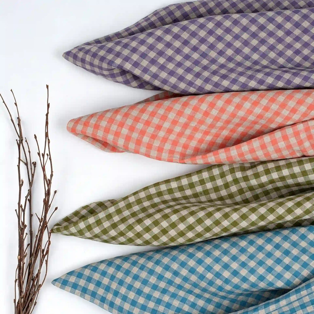 Four pieces of Longan Craft’s Colorful Checkered Linen Fabric By The Yard—100% washed linen gingham material in purple, pink, green, and blue—are arranged diagonally on a white surface beside a bundle of thin, bare twigs.