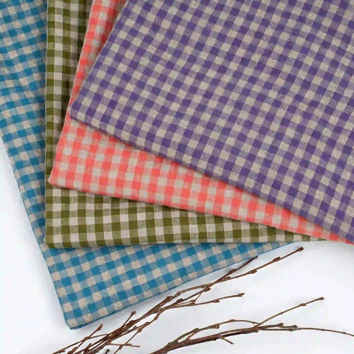 Four pieces of Longan Craft’s Colorful Checkered Linen Fabric By The Yard, featuring 100% washed linen gingham in blue, green, coral, and purple shades, are stacked in a fan shape on a white surface. Brown twigs rest nearby in the bottom left corner.