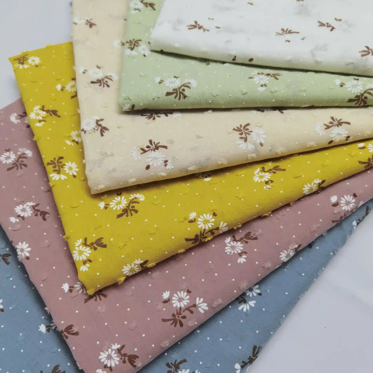 Five folded pieces of Longan Craft Colorful Daisy Print Swiss Dot Fabric—white, green, yellow, pink, and blue—feature small floral prints and dotted Swiss details on breathable 100% cotton.