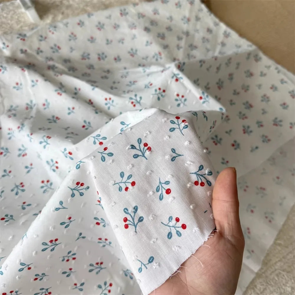 A hand holds Longan Craft’s Colorful Flower Printed Swiss Dot Fabric by the yard, a breathable cotton with a subtle dotted texture and floral pattern; more of this loungewear material is displayed in the background.