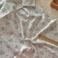 Longan Craft's Colorful Flower Printed Swiss Dot Fabric By The Yard, a breathable cotton with a floral pattern, is shown next to a wooden ruler and woven basket on a soft surface, highlighted by gentle natural light.