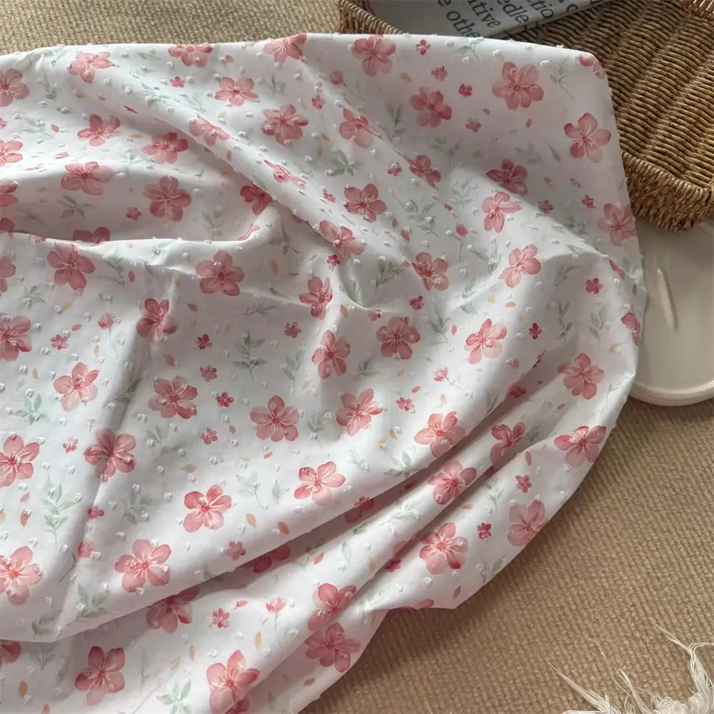 The Colorful Flower Printed Swiss Dot Fabric by Longan Craft, a cotton material with a vibrant floral pattern, is displayed draped over a beige mat beside a wicker basket and a white tray.