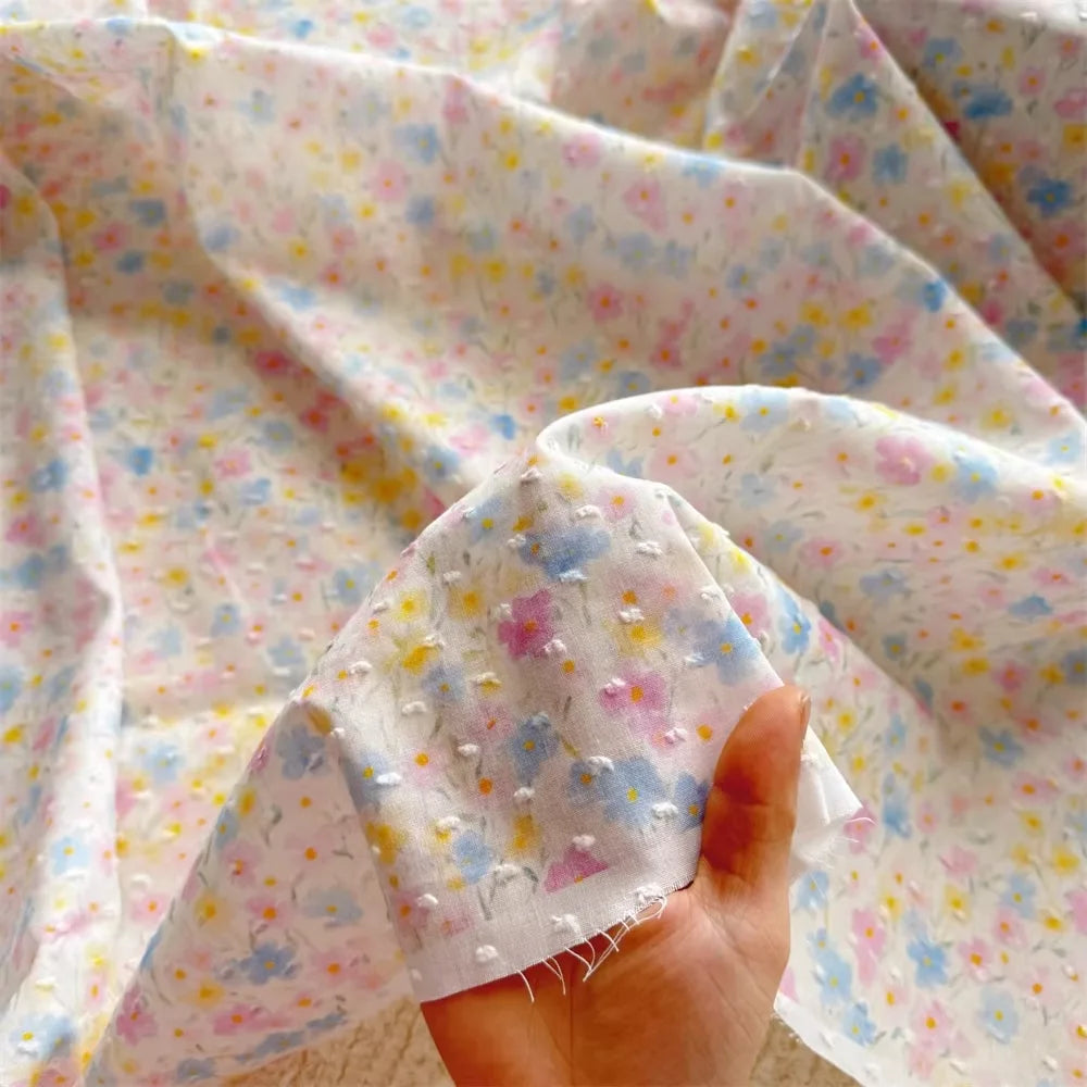 A hand holds Longan Craft’s Colorful Flower Printed Swiss Dot Fabric By The Yard—a soft, breathable, lightweight cotton with pastel floral patterns and dotted texture, ideal for loungewear. The material is sheer and slightly transparent.