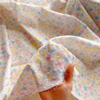 A hand holds Longan Craft’s Colorful Flower Printed Swiss Dot Fabric By The Yard—a soft, breathable, lightweight cotton with pastel floral patterns and dotted texture, ideal for loungewear. The material is sheer and slightly transparent.