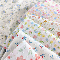 Several pieces of Longan Craft Colorful Flower Printed Swiss Dot Fabric by the yard, featuring blue, yellow, pink, and red floral patterns on cotton Swiss dot material, are neatly arranged on a floral background.