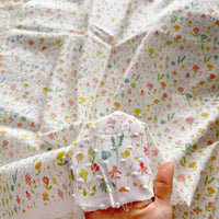 A hand holds a piece of Longan Craft's Colorful Flower Printed Swiss Dot Fabric by the yard, featuring a delicate cotton floral pattern that matches more of the same breathable material spread out in the background.