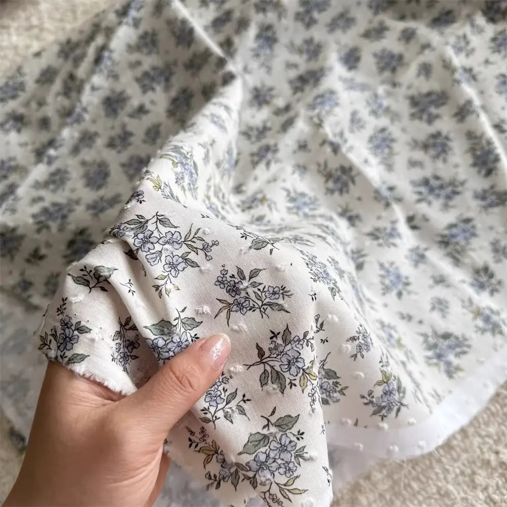A hand holds Colorful Flower Printed Swiss Dot Fabric by Longan Craft, a breathable cotton material with blue and green floral patterns, while more of this dotted Swiss fabric is spread out in the background.