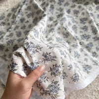 A hand holds Colorful Flower Printed Swiss Dot Fabric by Longan Craft, a breathable cotton material with blue and green floral patterns, while more of this dotted Swiss fabric is spread out in the background.