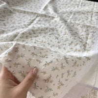 A hand displays Longan Craft's Colorful Flower Printed Swiss Dot Fabric by the Yard, a breathable cotton material featuring delicate yellow and green floral patterns—ideal for loungewear and shown on a subtly textured surface.