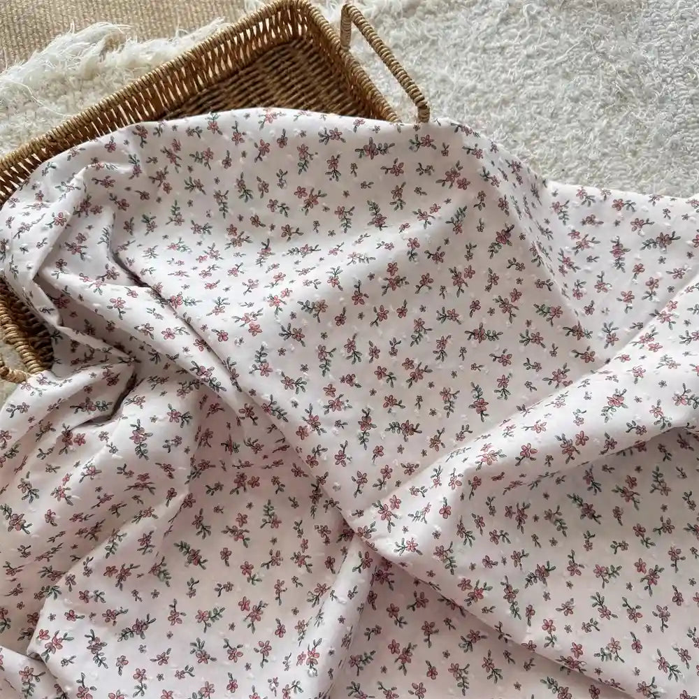 A wicker basket is partially covered by Longan Craft's Colorful Flower Printed Swiss Dot Fabric, a cotton material with a delicate floral pattern, resting on a textured white surface.