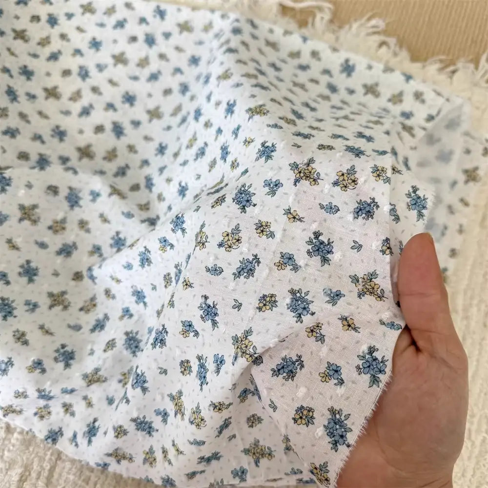 A hand holds Longan Craft’s Colorful Flower Printed Swiss Dot Fabric by the yard—breathable cotton with a blue and yellow floral pattern, frayed edges, and soft texture, perfect for loungewear.