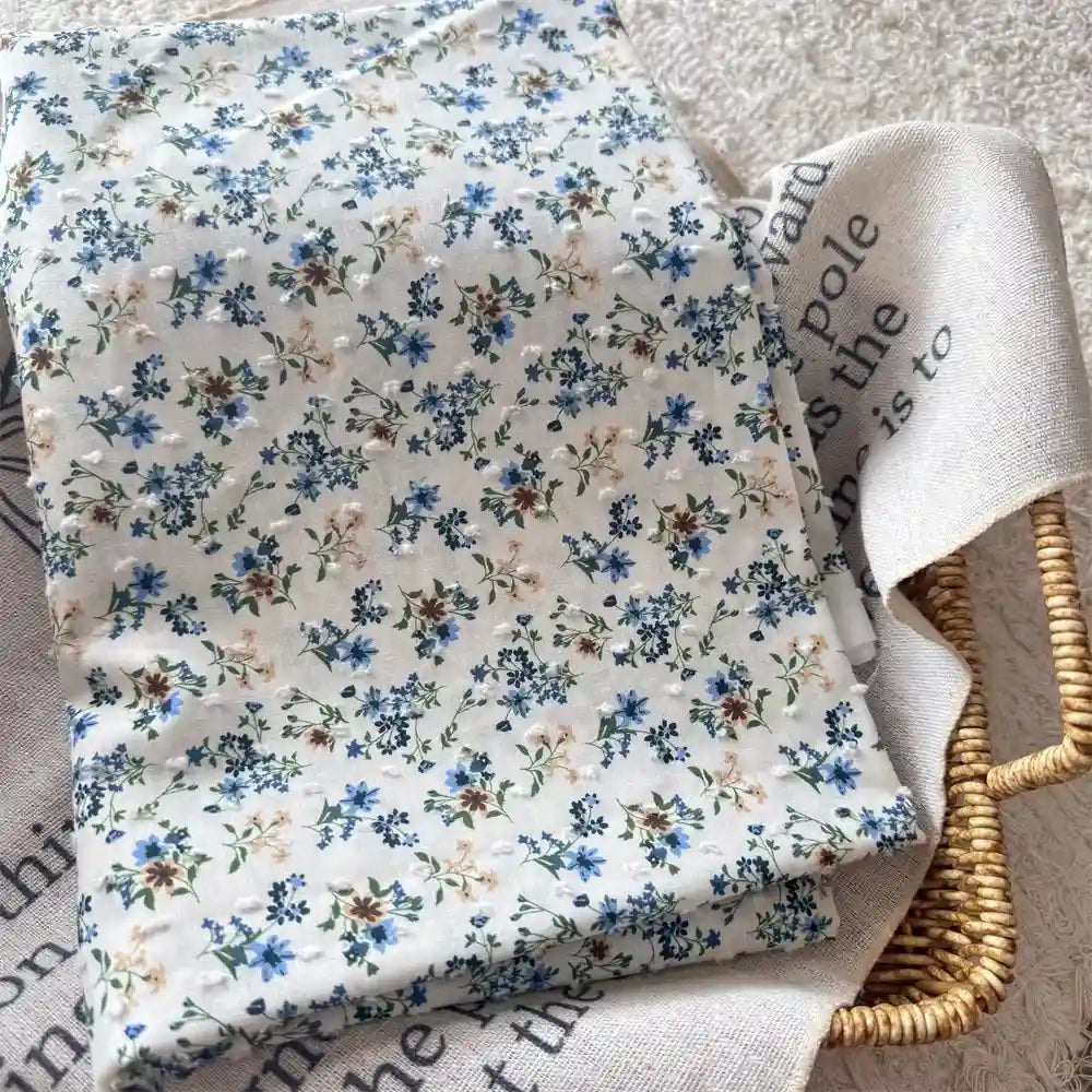 A folded piece of Longan Craft Colorful Flower Printed Swiss Dot Fabric by the Yard with a blue and beige floral cotton pattern rests atop another beige fabric with black text, both placed in a woven basket.