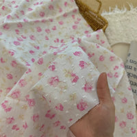 A hand holds Longan Craft’s Colorful Flower Printed Swiss Dot Fabric by the yard, a breathable cotton material with a pink and yellow floral pattern, ideal for loungewear. The background shows more of this dotted Swiss fabric and a woven basket.