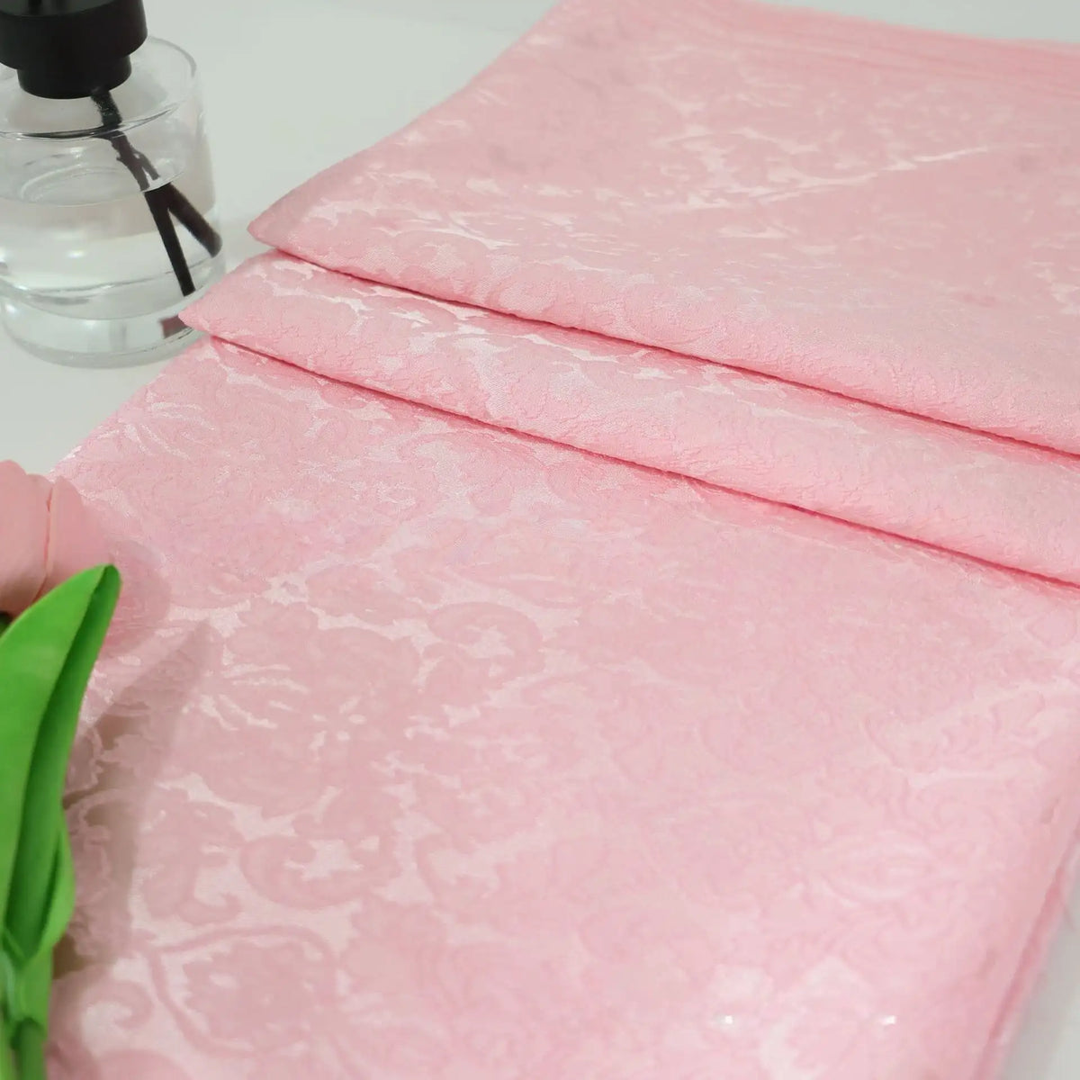 A folded piece of Colorful Jacquard Damask Fabric By The Yard from Longan Craft, featuring a floral pattern, lies on a white surface beside a green artificial flower and a clear spray bottle.