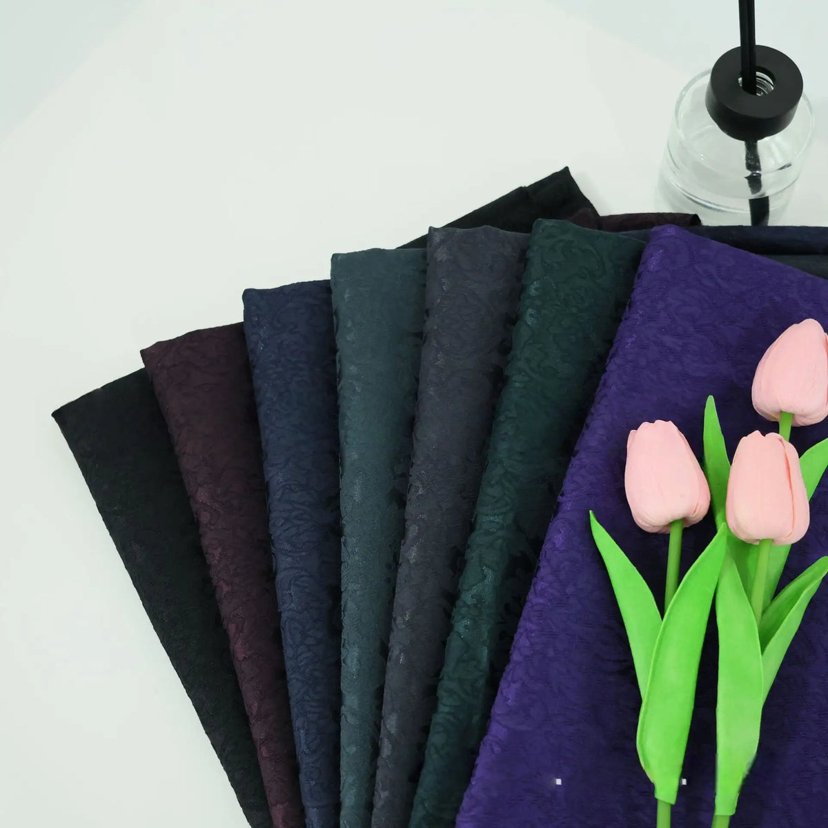 A display of Longan Craft's Colorful Jacquard Damask Fabric By The Yard—featuring a floral pattern in black, blue, green, and purple—rests beside pink artificial tulips and a clear vase, ideal for creative home décor.