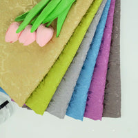 A bouquet of pink tulips rests atop seven neatly stacked Colorful Jacquard Damask Fabrics by Longan Craft, with elegant floral patterns in gold, green, gray, blue, purple, and brown fanned out on a white surface.