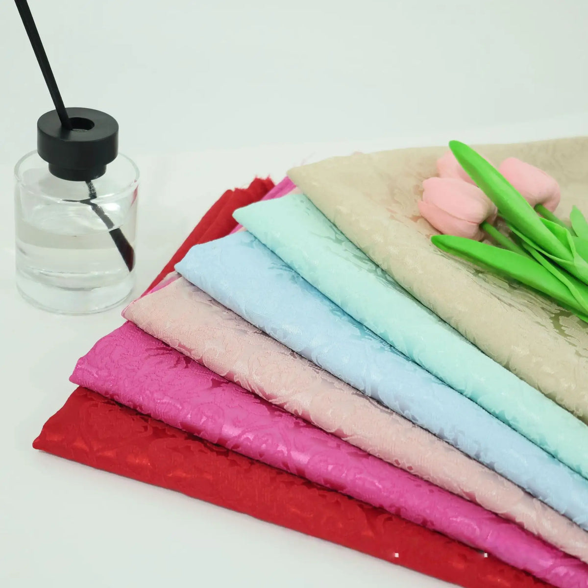 A stack of Colorful Jacquard Damask Fabric By The Yard from Longan Craft, in red, pink, blue, and beige floral patterns, rests on a white surface beside a glass jar and pink tulips—ideal for creative home décor projects.