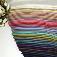 A neatly arranged stack of Longan Craft’s Colorful Lightweight Ramie Fabric By The Yard is displayed in an array of solid hues, from crisp white and gentle pastels to vivid and deep tones. The pure, soft ramie material lends an airy feel to the presentation, while artificial white roses placed at the side on a clean white surface enhance the vibrant and inviting display—perfect for garment making.
