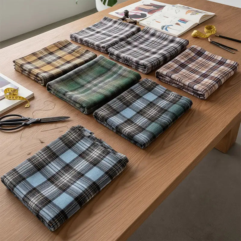 Seven folded pieces of Longan Craft Colorful One Side Sueded Flannel Fabric by the Yard, featuring a variety of yarn-dyed cotton plaid designs, are neatly arranged on a wooden table alongside scissors, a measuring tape, and other sewing tools.