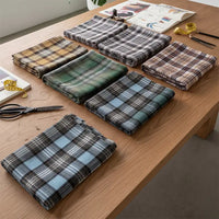 Seven folded pieces of Longan Craft Colorful One Side Sueded Flannel Fabric by the Yard, featuring a variety of yarn-dyed cotton plaid designs, are neatly arranged on a wooden table alongside scissors, a measuring tape, and other sewing tools.