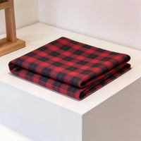 A neatly folded piece of Longan Craft Colorful One Side Sueded Flannel Fabric By The Yard—featuring a vibrant red and black yarn-dyed cotton plaid—is displayed on a white surface beside a wooden planter.