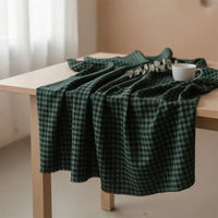 A piece of the Colorful One Side Sueded Flannel Fabric By The Yard from Longan Craft, a yarn dyed cotton plaid flannel with a green and black checkered pattern, is draped over a light wooden table, topped with a white cup and a eucalyptus branch.