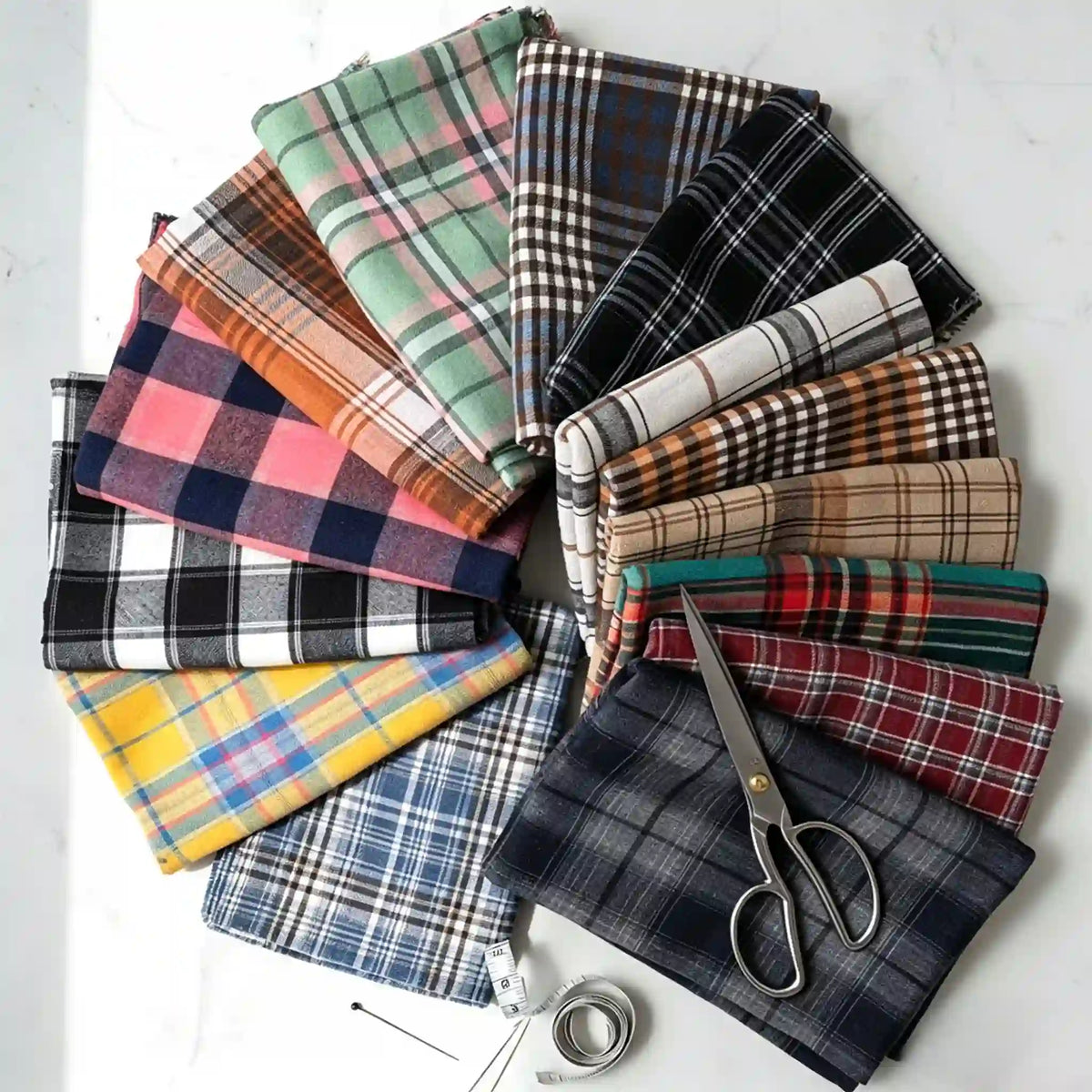 A selection of vibrant Longan Craft Colorful One Side Sueded Flannel Fabric By The Yard, featuring yarn-dyed cotton plaid designs from the Super Snuggle Fall Flannel collection, is displayed in a circular arrangement with scissors, a tape measure, and pins on a white surface.