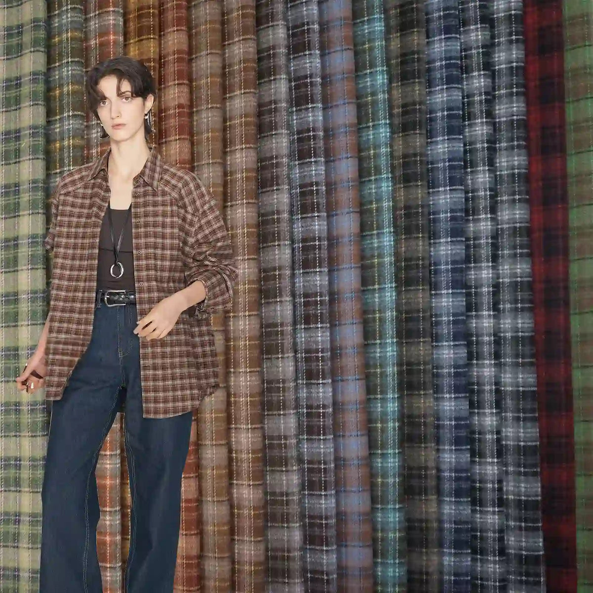A person in a brown plaid shirt and jeans stands before a display of Longan Craft's Colorful Polyester Rayon Mammoth Flannel Fabric by the yard—plaid brushed flannelette in brown, blue, gray, and green shades for apparel.