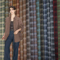 A person in a brown plaid shirt and jeans stands before a display of Longan Craft's Colorful Polyester Rayon Mammoth Flannel Fabric by the yard—plaid brushed flannelette in brown, blue, gray, and green shades for apparel.