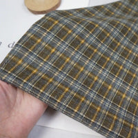 A hand holds Longan Craft's Colorful Polyester Rayon Mammoth Flannel Fabric By The Yard, featuring a snuggly gray, blue, and yellow plaid pattern. The soft, brushed flannelette fabric is shown with a round wooden object in the background.