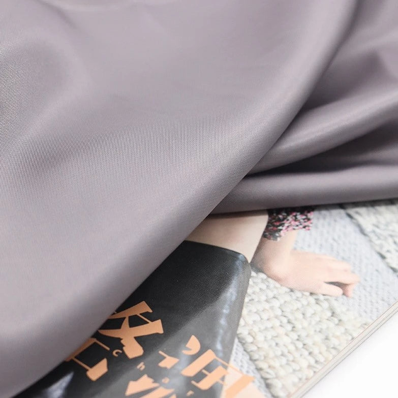 A swatch of Longan Craft’s Colorful Satin Viscose Fabric with twill weave in light gray drapes over a magazine page, partially obscuring an image of legs on a rug, with Japanese text visible at the bottom.